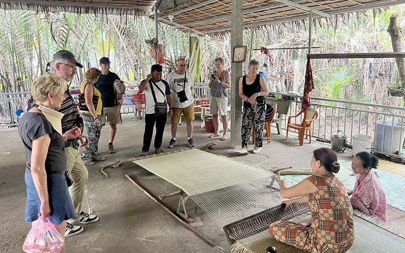 Khách du lịch nước ngoài theo dõi công đoạn dệt chiếu tại xã Nhơn Thạnh (thành phố Bến Tre, tỉnh Bến Tre).